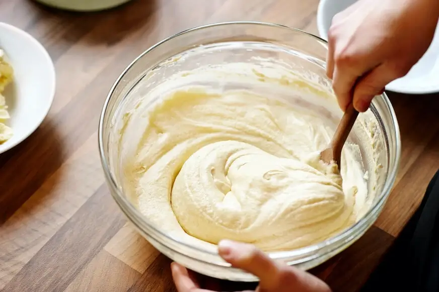 Person mixing cake batter in a glass bowl with a wooden spoon.