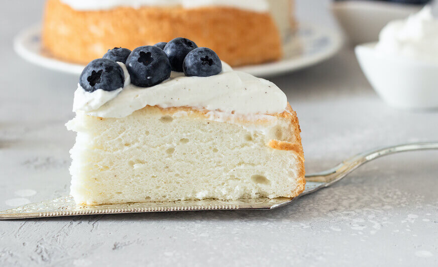 Slice of angel food cake topped with whipped cream and blueberries, showing its light, fluffy texture.