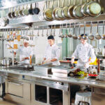 A commercial kitchen with kitchen staff at work. A number of pots and pans are hanging, and other ingredients and tools are neatly lined up.