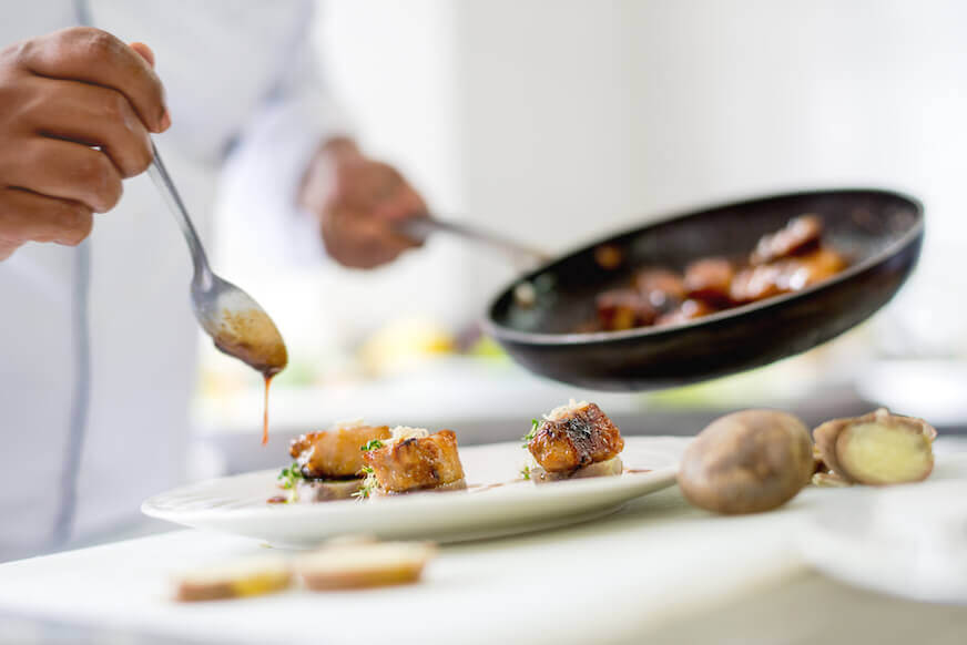 Chef holding a pan while drizzling sauce on a plate with a spoon