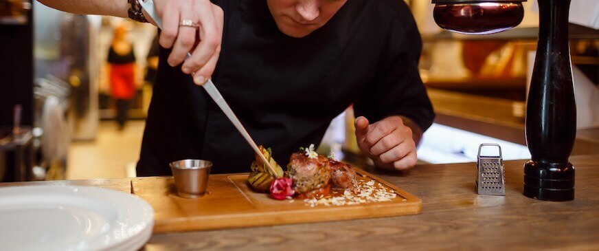 Chef in a black uniform works on a meat dish on a wooden plate