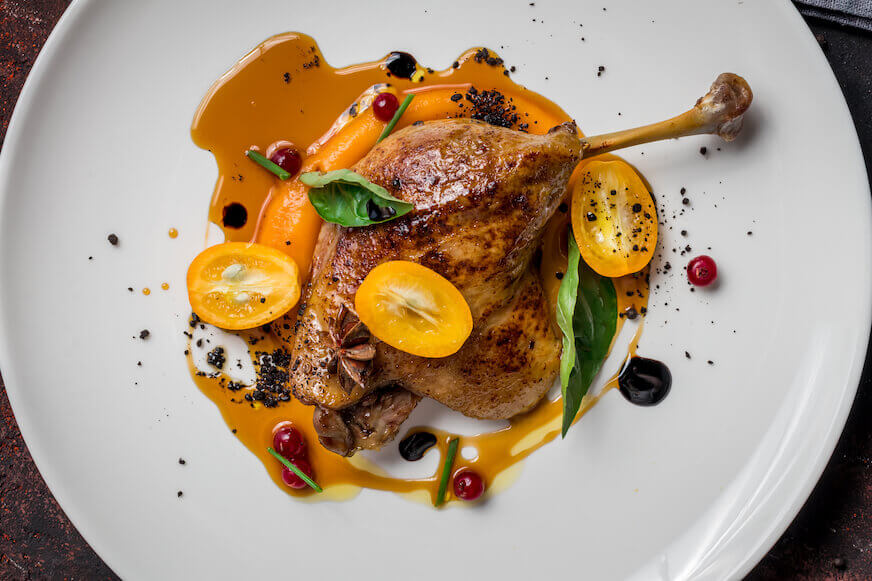 Duck leg confit on a white plate covered in a sauce and tomatoes