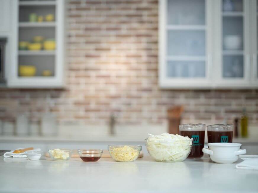 Mise en place for French onion soup, with dark brown broth, a bowl of sliced onions, cheese, worcestershire sauce, butter, and salt.