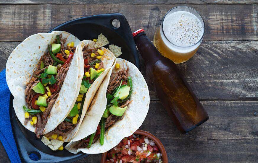 Three tacos on a skillet next to a bottle and a glass of beer on a wooden table