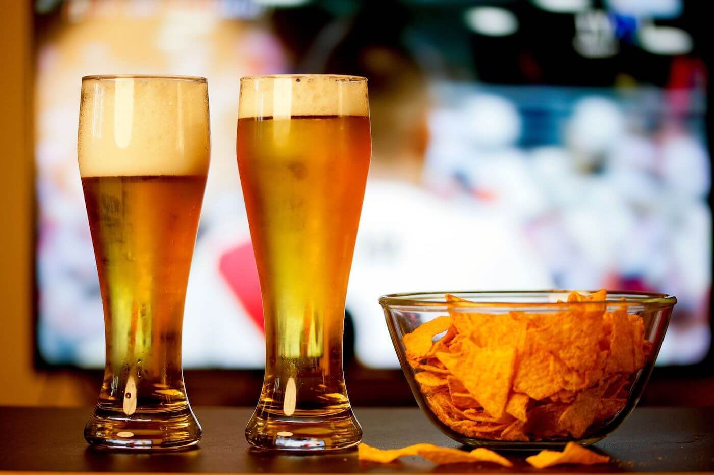 Two tall glasses of beer next to a bowl of chips
