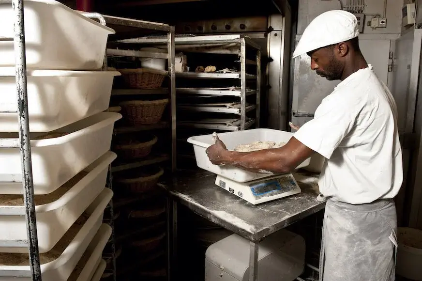 A baker in a white uniform and cap weighing dough in a commercial kitchen, surrounded by industrial baking racks.