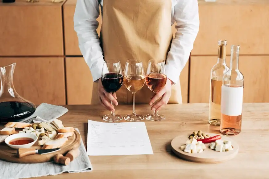 A person in an apron holding three wine glasses with different types of wine, with a table set with cheese, bread, olives, and a wine tasting sheet.