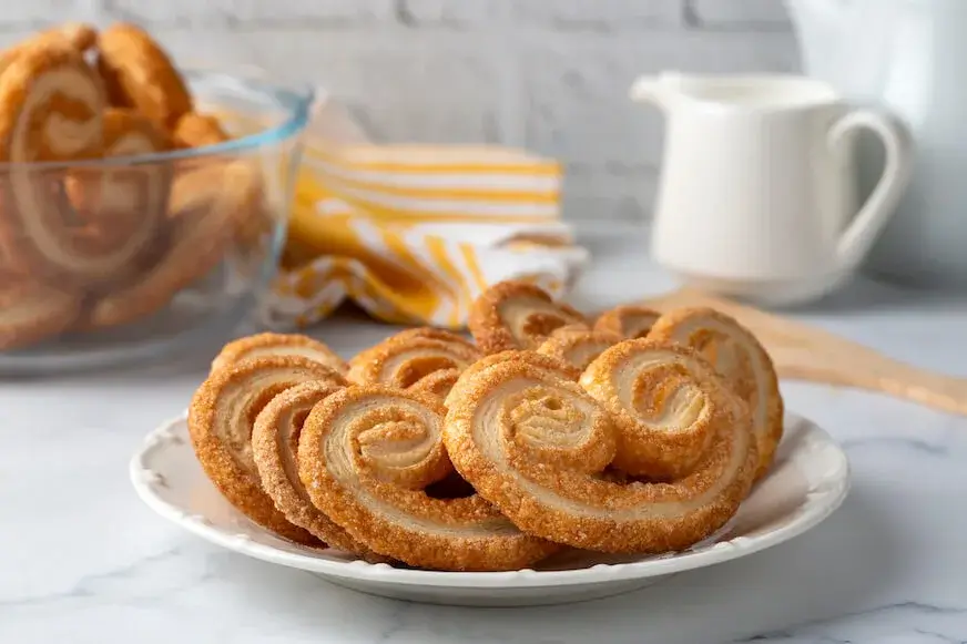 A plate of palmiers sits on a white plate atop a marble countertop.