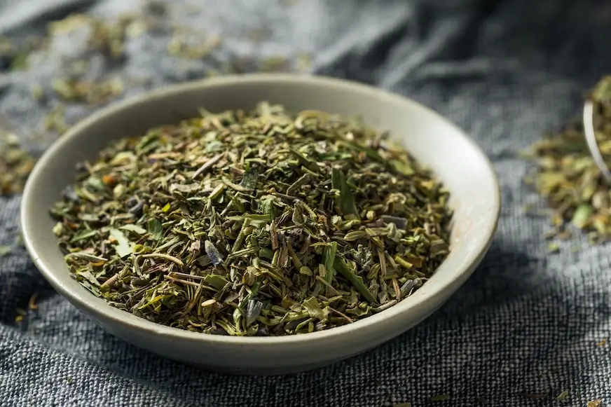 A shallow bowl filled with a mix of dried herbs, sitting on a rustic blue-gray fabric surface.