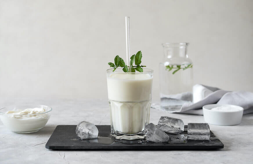 A tall, clear glass of milk sits centered on a marble countertop with ice cubes and a short white dish of yogurt beside it.