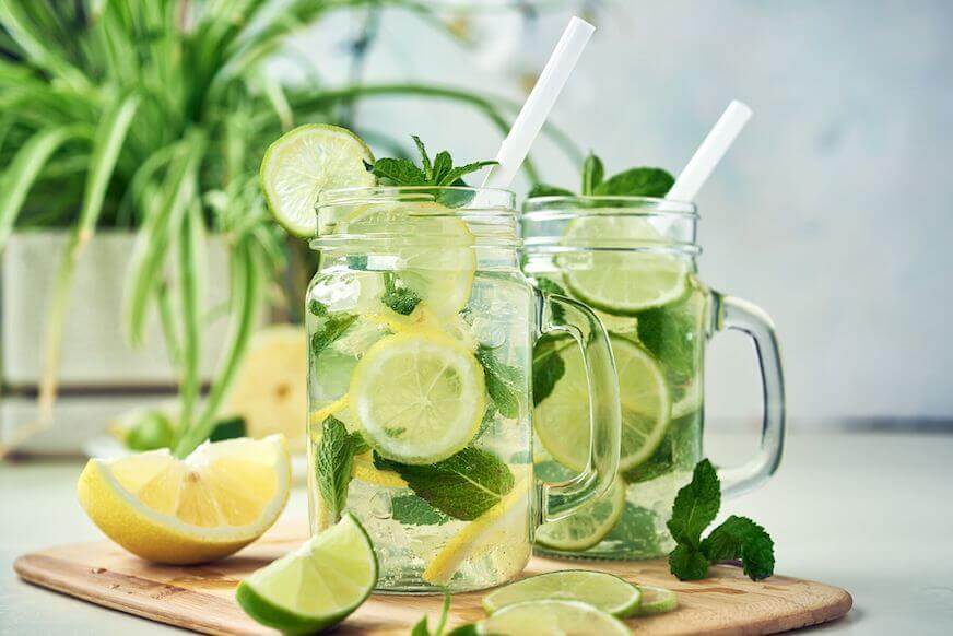 Two mason jar glasses with homemade lemon, lime, and mint infused water sit on a wooden dining table.