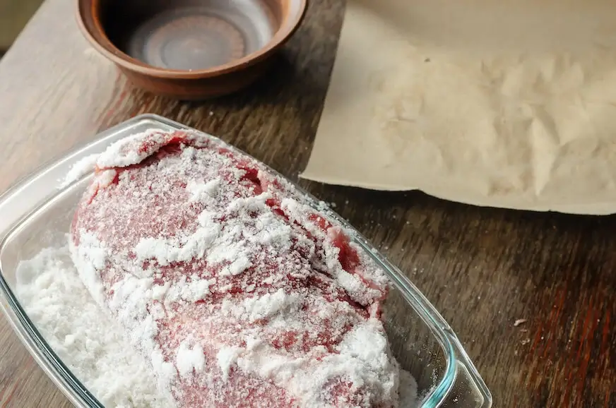 A pork loin is covered in salt and sits in a glass container, ready for the next steps in dry curing.
