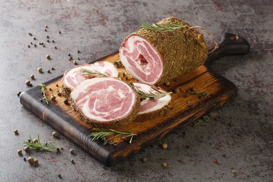 Pancetta covered in salt, herbs, and spices sits on a wooden cutting board.