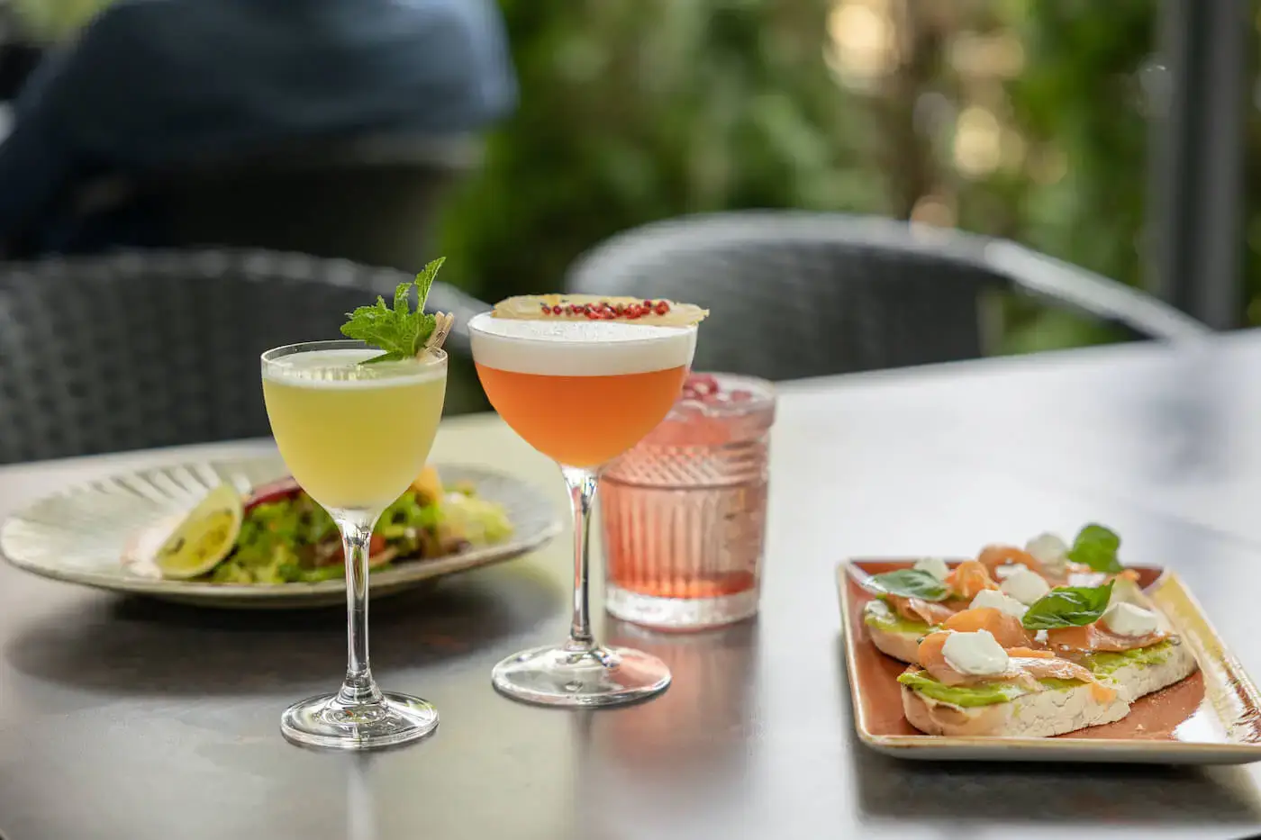 Colorful cocktails and mocktails in various glassware paired with two plates of food on a table.