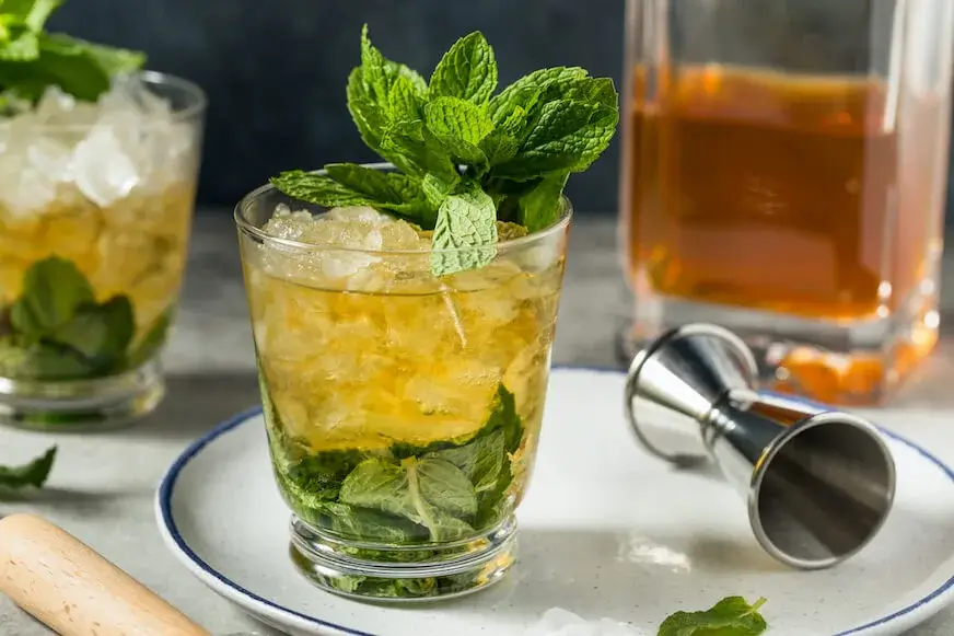 Mint julep in a glass with crushed ice and fresh mint leaves, bourbon bottle in background.
