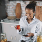 A woman is smiling at her phone while standing in the kitchen holding an apple