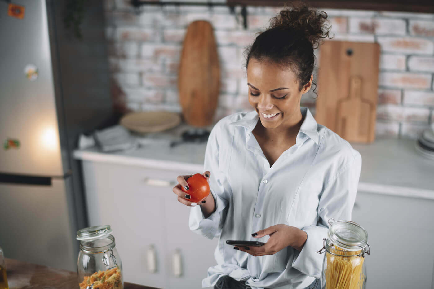 A woman is smiling at her phone while standing in the kitchen holding an apple