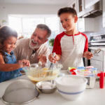 Brother and sister standing at the kitchen table making cake mix with their grandfather, close up