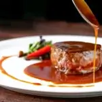 A spoon pours glossy brown demi-glace over a steak, showing the deep color and texture of Espagnole-based sauce.