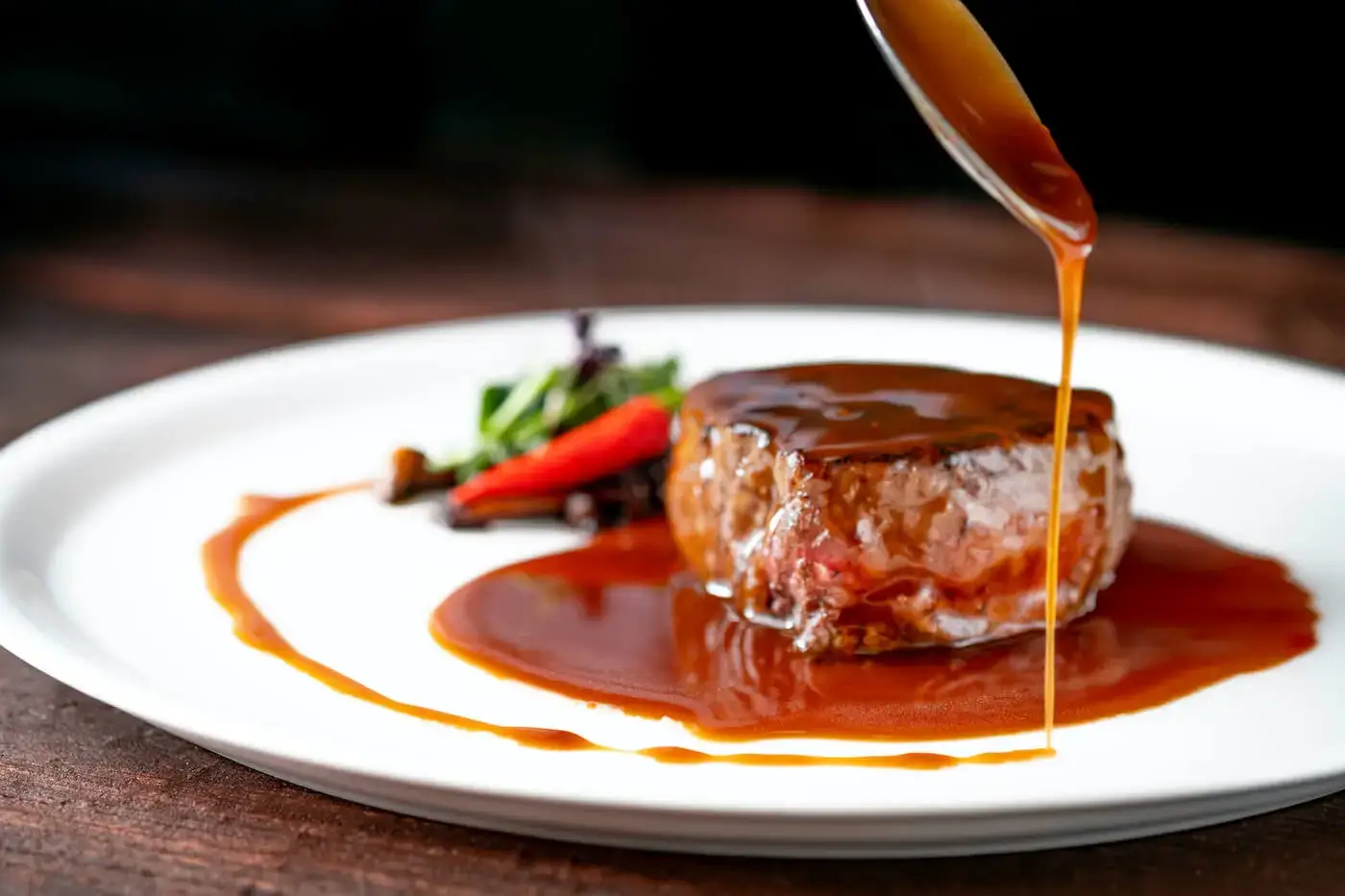 A spoon pours glossy brown demi-glace over a steak, showing the deep color and texture of Espagnole-based sauce.