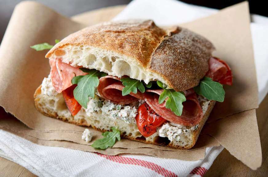 A closeup of a sandwich made with ciabatta bread and filled with salami, arugula, and red peppers.