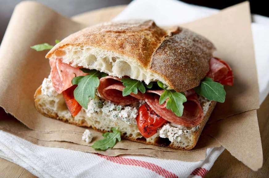Tampilan jarak dekat dari sandwich yang dibuat dengan roti ciabatta dan diisi dengan salami, arugula, dan paprika merah.