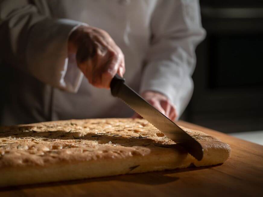 A hand holds a bread knife and slices into a sheet of fresh focaccia bread on a brown cutting board.