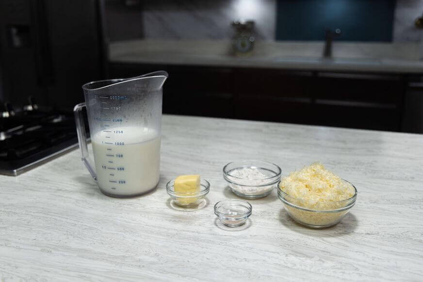 Five ingredients sit on a countertop: milk in a measuring cup, then butter, flour, pepper, and grated cheese, each in pinch bowls.