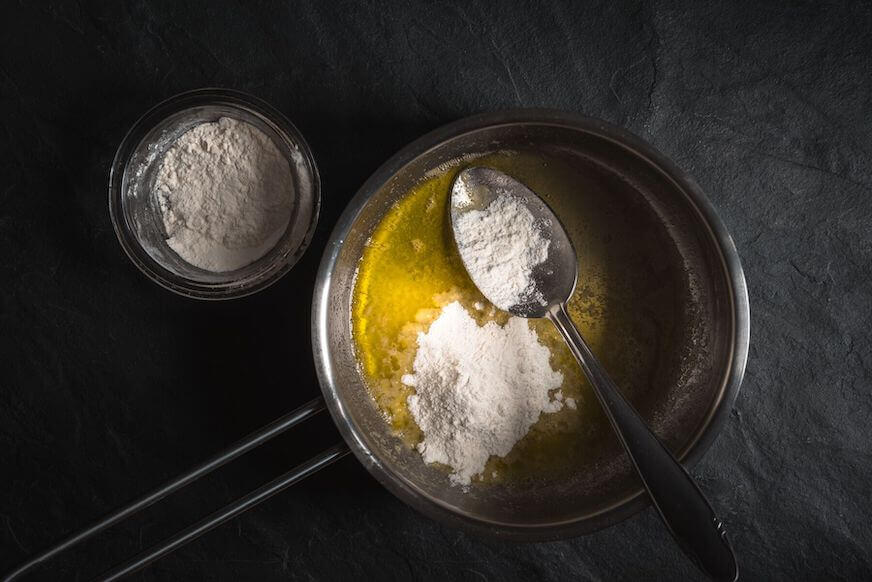 Overhead view of a pan with melted butter and a spoon with flour, resting on the pan. Another spoonful of flour is already in the butter, but not mixed yet.